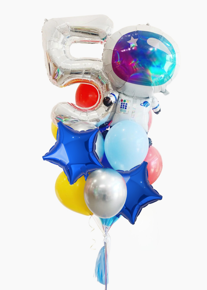 A cluster of birthday balloons on a plain white background, including a large metallic silver balloon shaped like the number 5, a round foil balloon with a colorful galaxy space design, a small astronaut balloon, blue star balloons, a light blue balloon, a yellow balloon, a silver balloon, and a pink balloon.