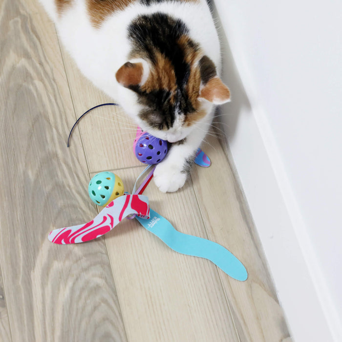 A white and brown cat plays on a light wooden floor with Quirky Kitty Catnip Seaweed Tumblers 2pk—ocean-themed cat toys with catnip—and colorful balls, next to a white wall.
