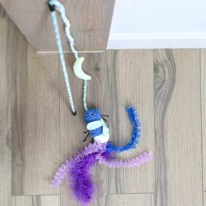 The Quirky Kitty Firefly Wand, a glowing cat wand toy with catnip and silvervine, hangs from a rope on wood. It features blue and white colors, purple and lavender ribbons, a crescent moon accent, and is set against light wood and a white wall.