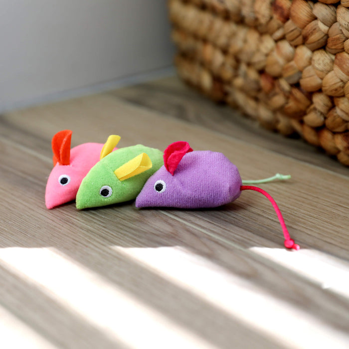 Three Quirky Kitty Glow Mice—catnip & silvervine-filled cat toys in pink, green, and purple—sit on the floor near a basket as sunlight streams in, perfect for interactive nighttime play.