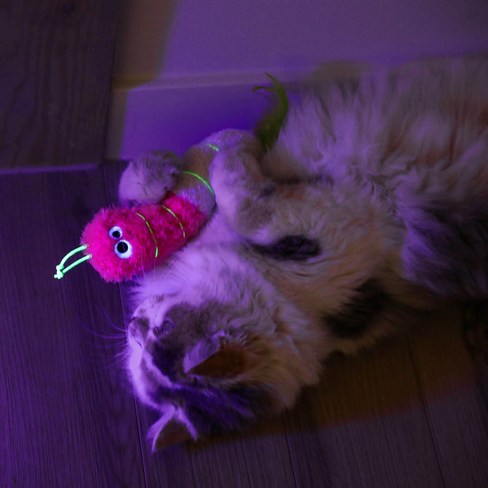 A fluffy cat lies on a wooden floor, hugging the Quirky Kitty Glow Worm Kicker by Quirky Kitty—a catnip and silvervine toy that glows at night—under purple lighting.