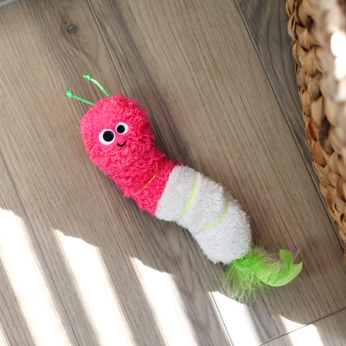The Quirky Kitty Glow Worm Kicker—a catnip and silvervine toy that glows at night—lies on a wooden floor near a woven basket, sunlight casting playful shadows nearby.