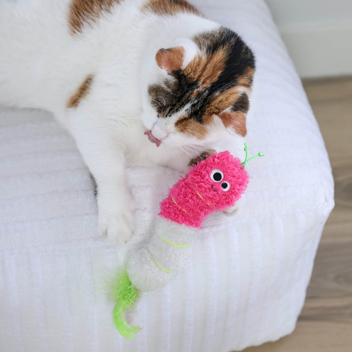 A white, brown, and black cat is lying on a white textured surface licking its paw next to the Quirky Kitty Glow Worm Kicker—a pink and white catnip and silvervine toy by Quirky Kitty that glows at night with playful details.