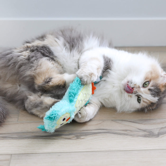 A fluffy cat with white, gray, and tan fur lies on a wooden floor, playfully biting the Quirky Kitty Night Owl Kicker—a glow-in-the-dark catnip and silvervine toy—while sticking its tongue out.