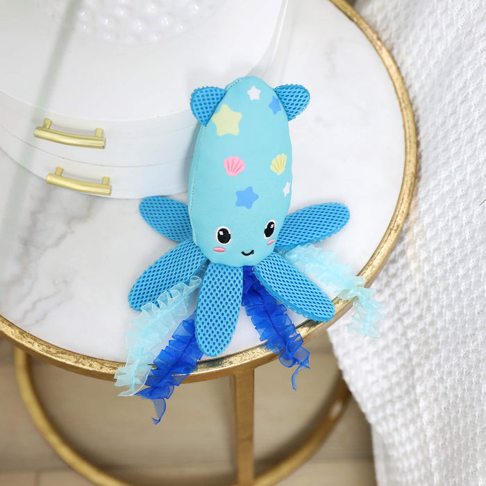 The Quirky Kitty Octopurr Kicker, an ocean-themed catnip toy, features star and shell patterns, mesh fins, and ruffled blue tentacles. It sits on a gold-rimmed marble side table beside a white textured blanket.